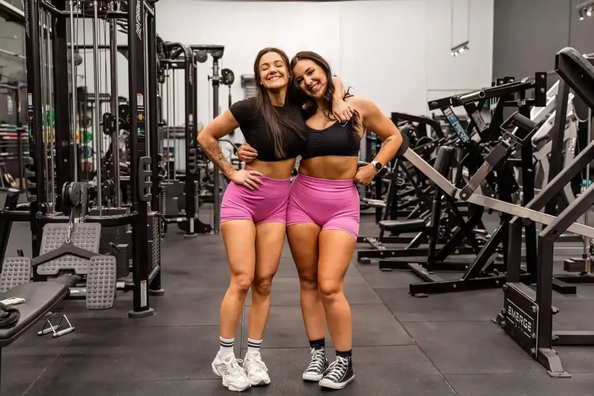 Two women in black tops and pink shorts posing together among gym equipment